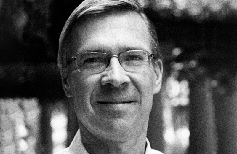 A black and white headshot of a person wearing glasses, smiling, with blurred outdoor background.