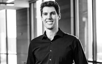 A person smiling in a professional headshot, wearing a dark shirt, with a background of large windows and a softly lit interior.