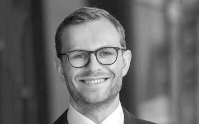 A person wearing glasses and a suit, smiling against a blurred background.